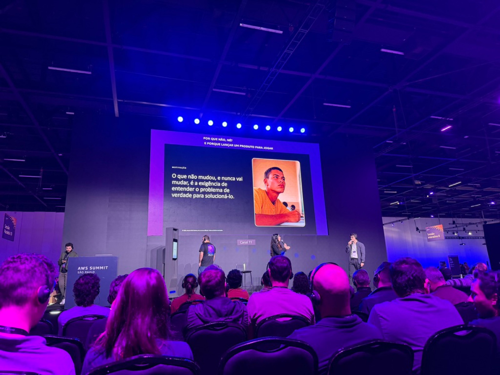 Telão de uma sessão do AWS Summit São Paulo exibindo a frase citada, com o palco e a plateia em primeiro plano